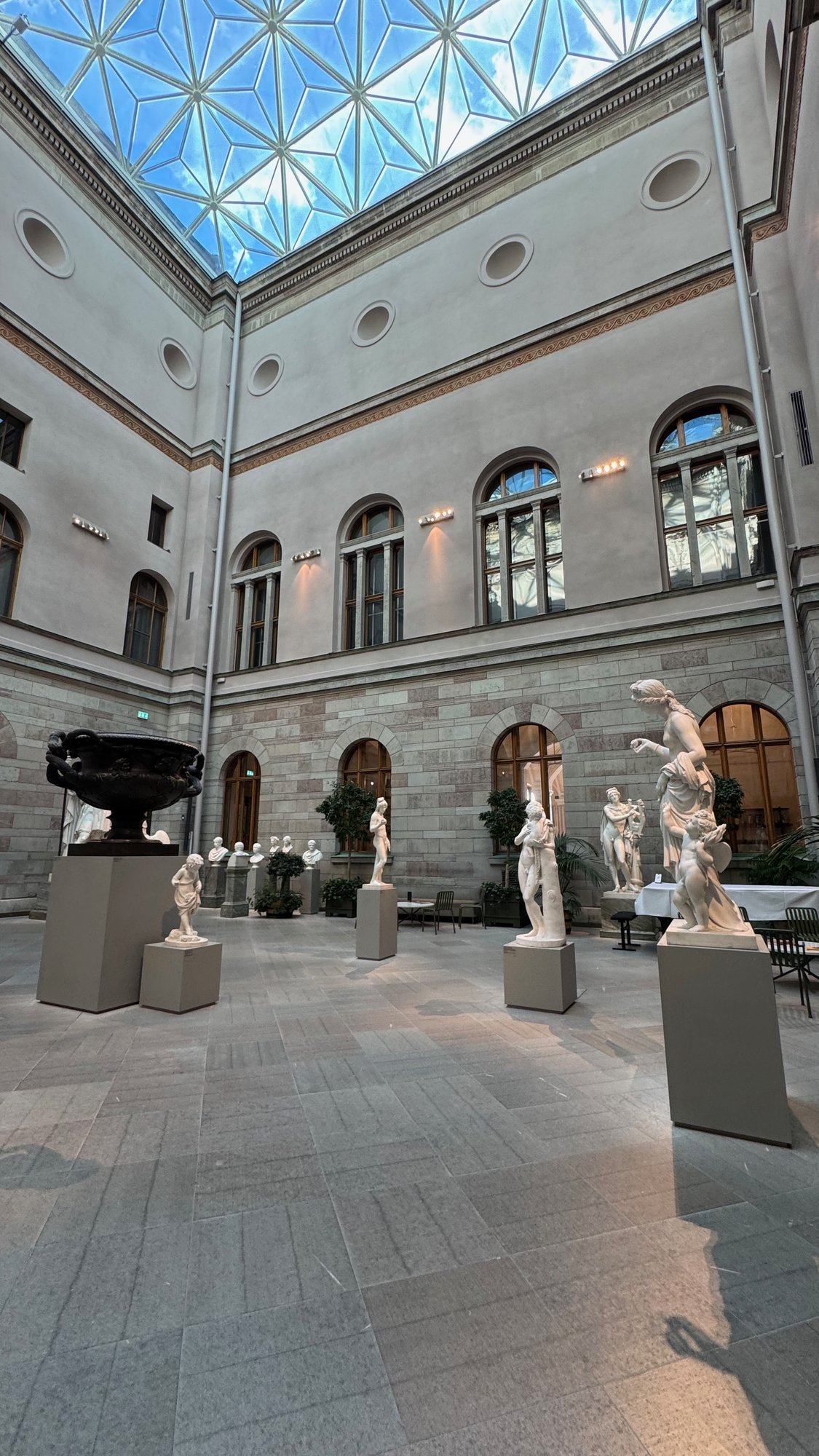 Sculpture courtyard with a star-patterned glass roof, white marble statues on gray bases, a large dark bronze vase on the left, café tables on the right and arched windows in a stone wall.