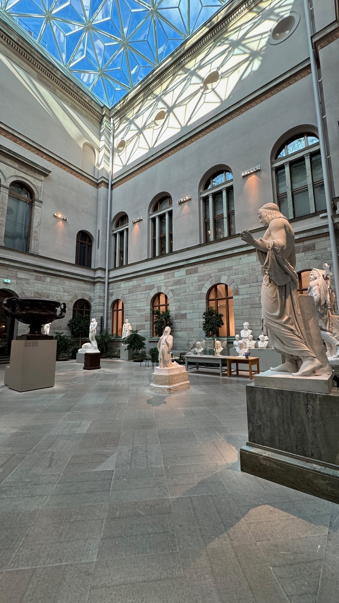 Sculpture courtyard with a star-patterned glass roof, a white marble figure with a raised hand on a gray base in front, more statues and busts behind, and a dark bronze vase on the left.