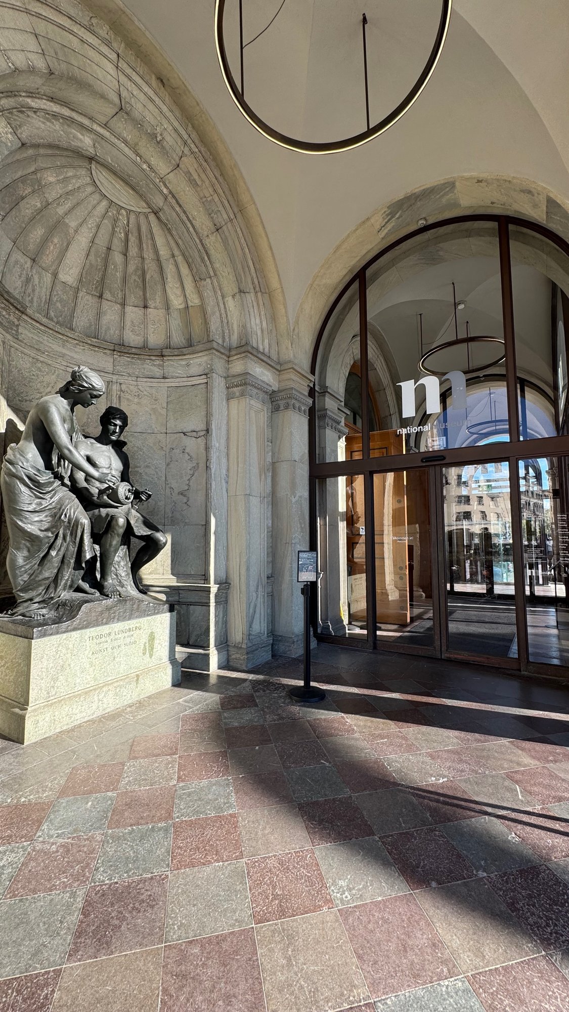 Bronze statue group of a woman and a seated man holding a round object, in a stone niche beside the glass entrance doors, with the names Teodor Lundberg and Konst och slöjd on the base.