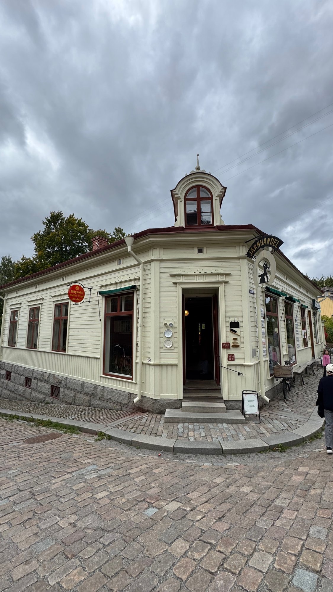Cream wooden corner house with a small tower on the roof, a black Järnhandel sign, an open door and a bicycle in the window, on a cobbled street under a cloudy sky.