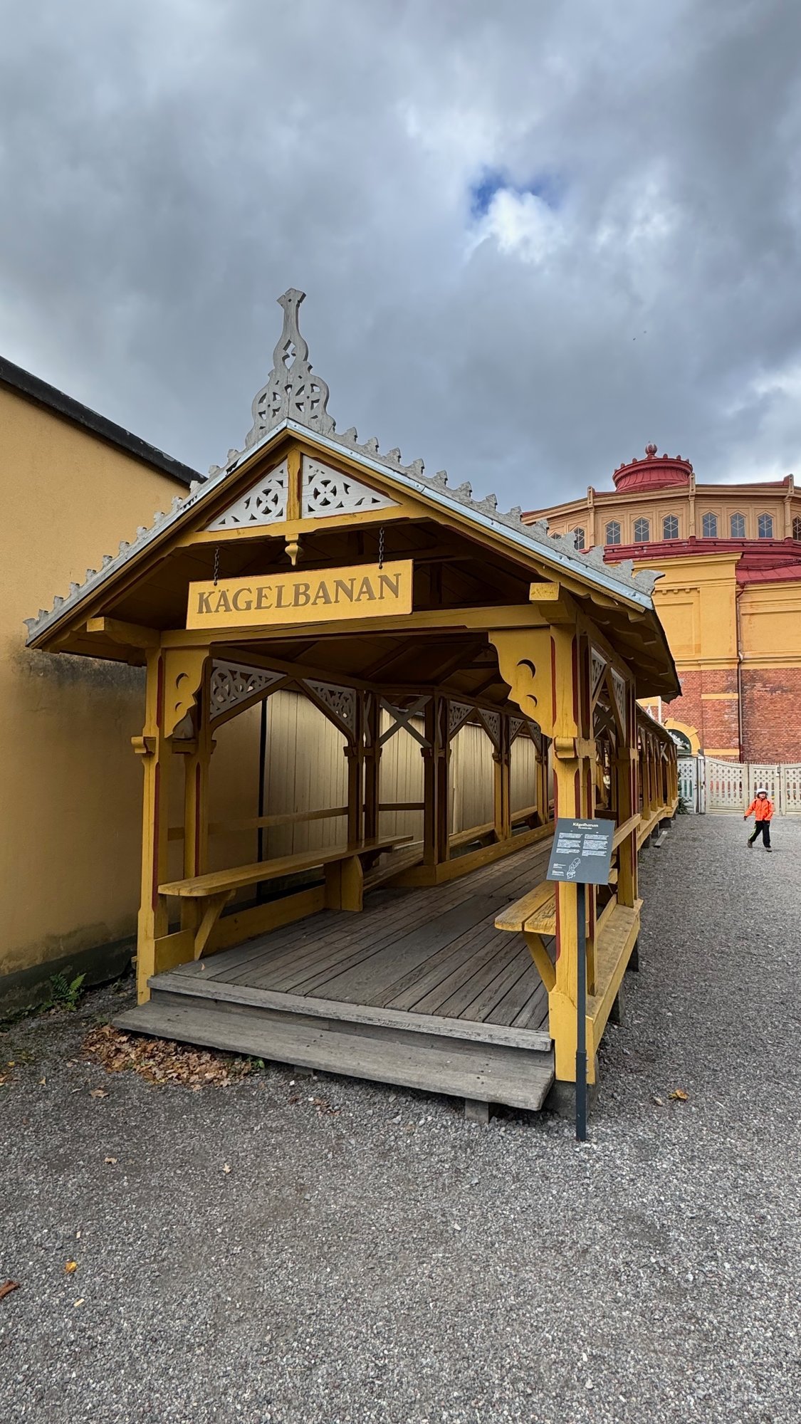 Yellow wooden pavilion with carved gable ornaments and the sign Kägelbanan, open on the sides with wooden benches, beside a gravel path, with a yellow and brick building behind.