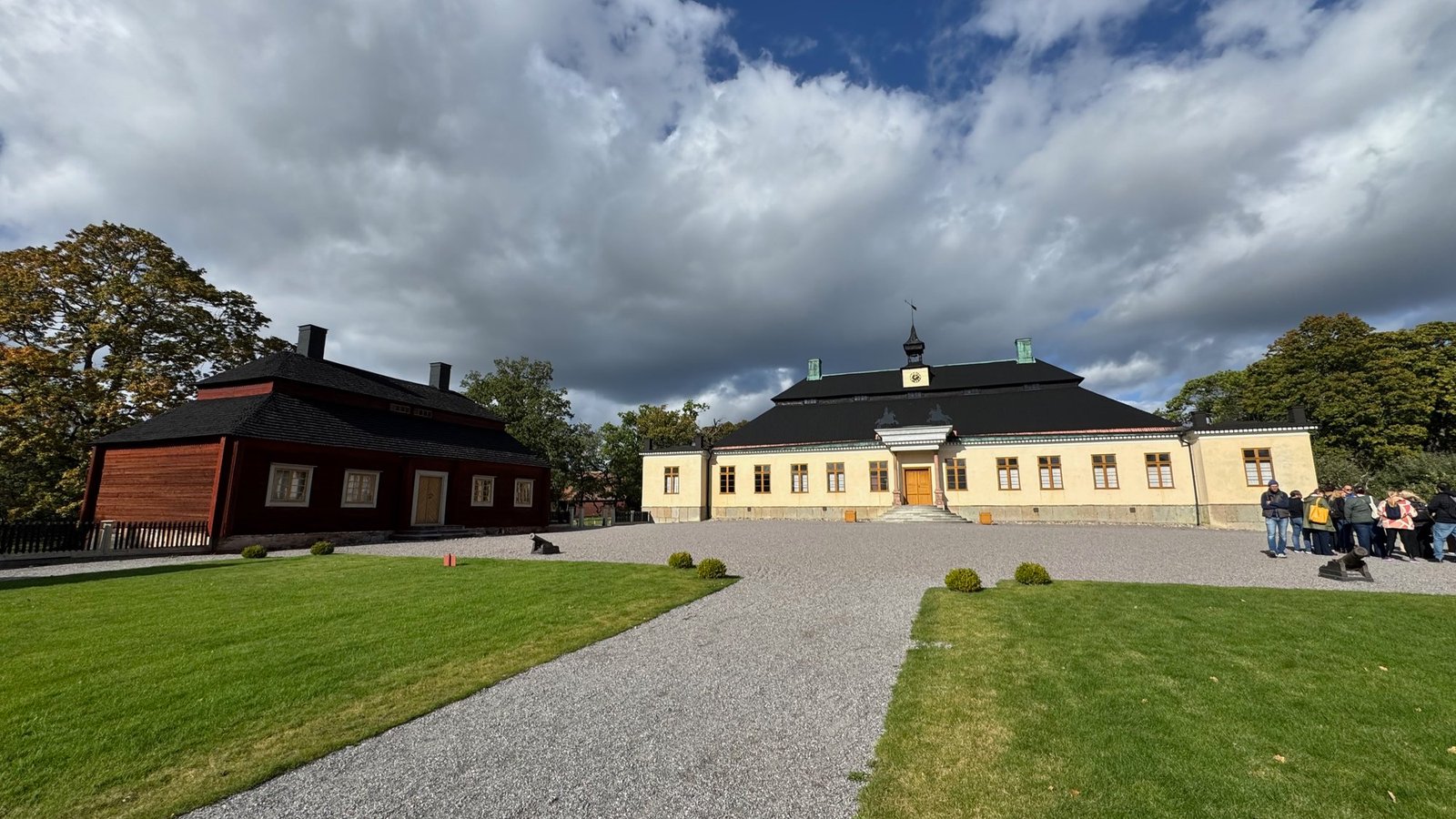 Long cream building with a black roof and a small clock tower beside a dark red wooden building, on a gravel yard with lawn and two small cannons, under a cloudy sky.