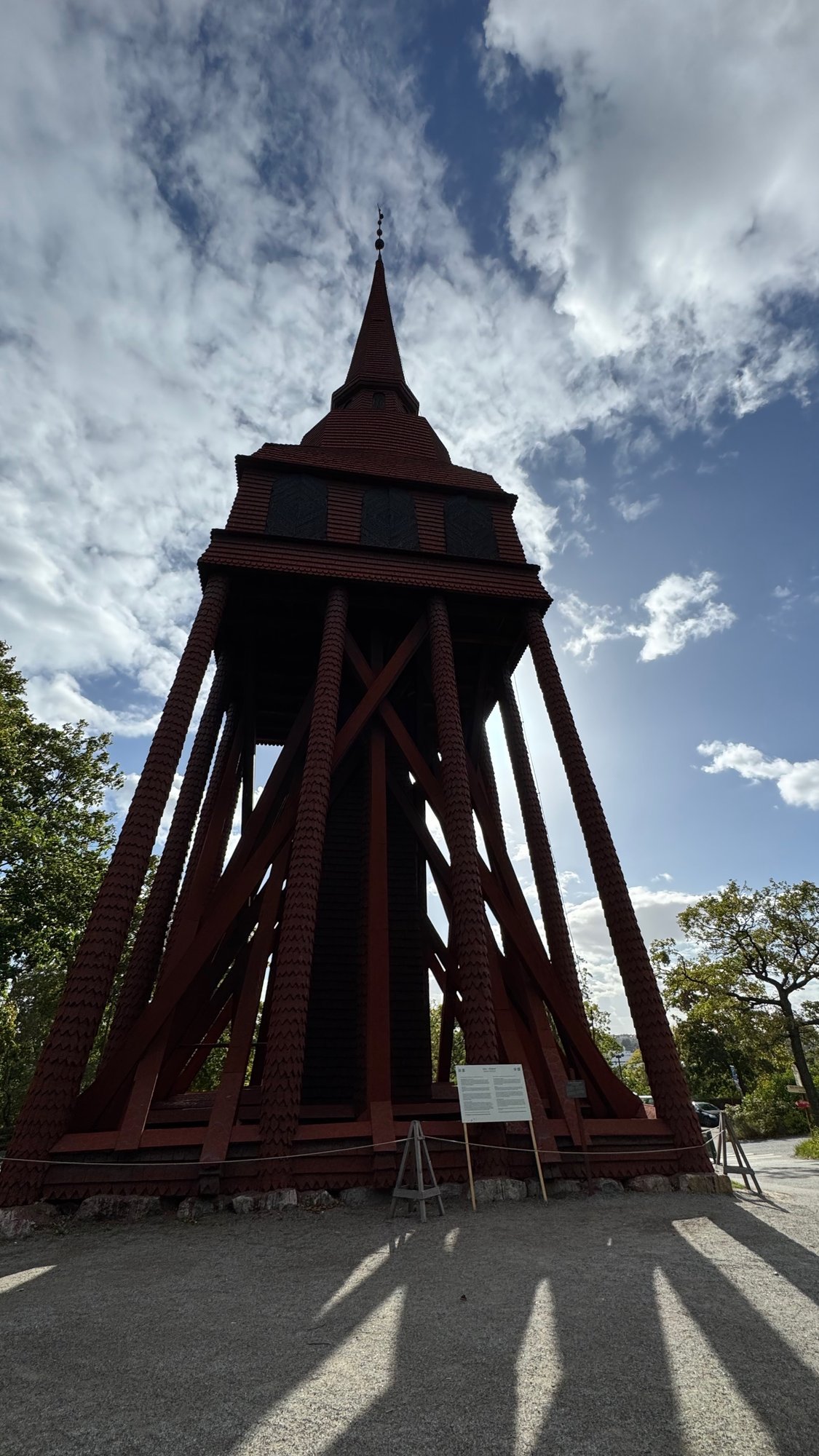 Tall red wooden bell tower seen from below, with slanting legs, cross bracing, shingled sides and a thin spire against a blue sky with clouds, and an info sign at its base.