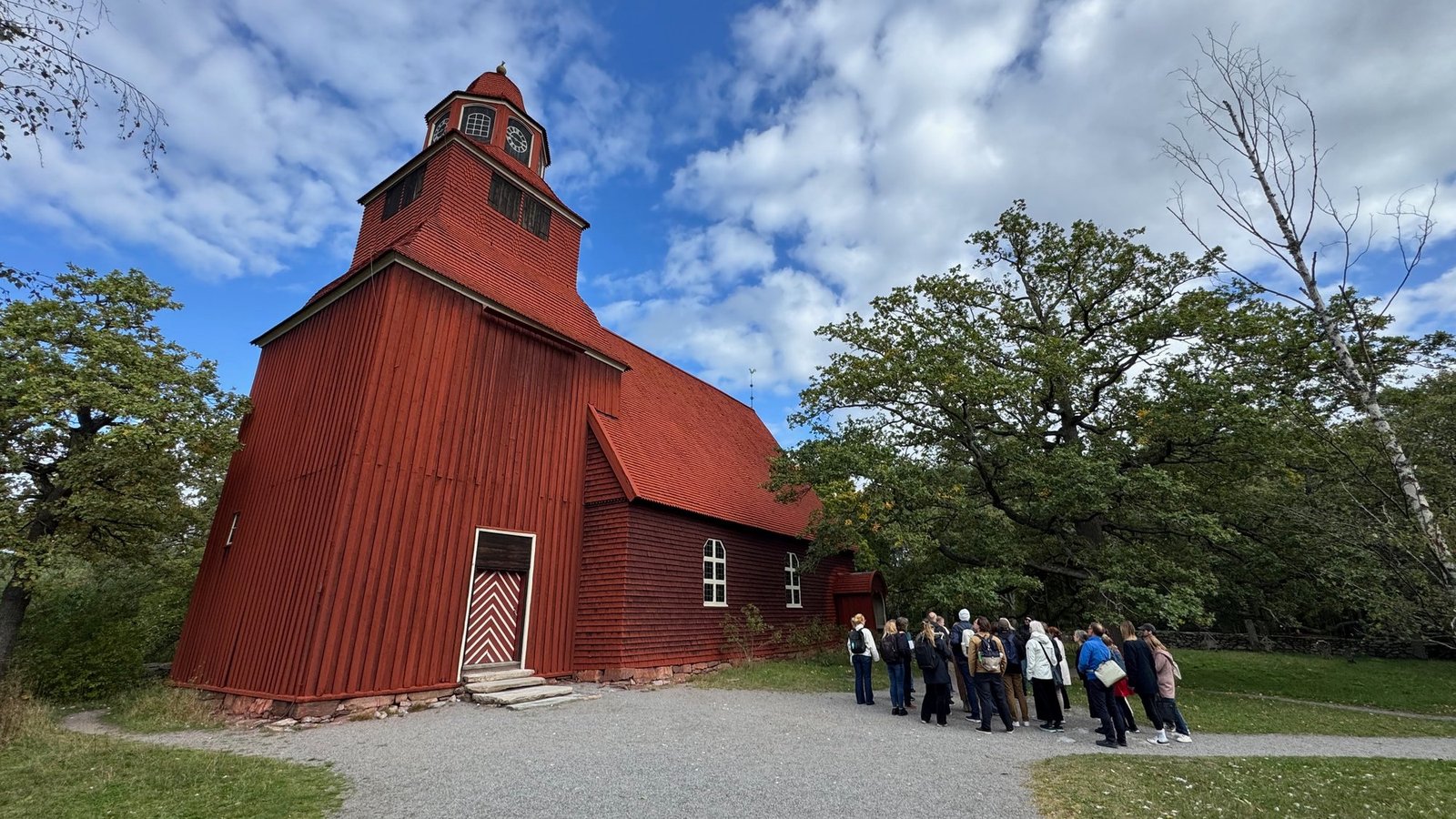 Large red wooden church with a tall clock tower, a white and red zigzag pattern on the door, green trees and a group of visitors by the wall, under a blue sky with white clouds.