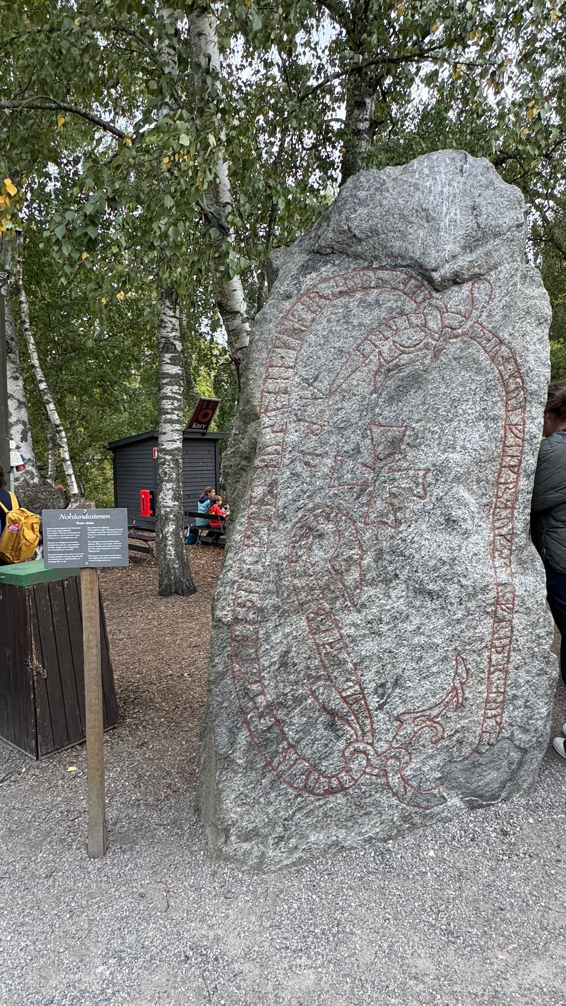 Tall gray rune stone with red painted runes, a snake band and a cross, with the sign Norslundastenen at its foot and birch trees and a small kiosk behind.