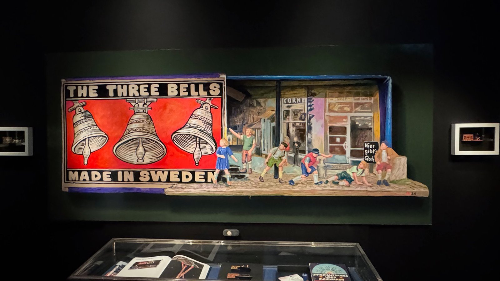 Matchbox art on a dark wall: a red label with the text The Three Bells Made in Sweden and an open drawer with a painted street scene and children playing, above a glass case with books.