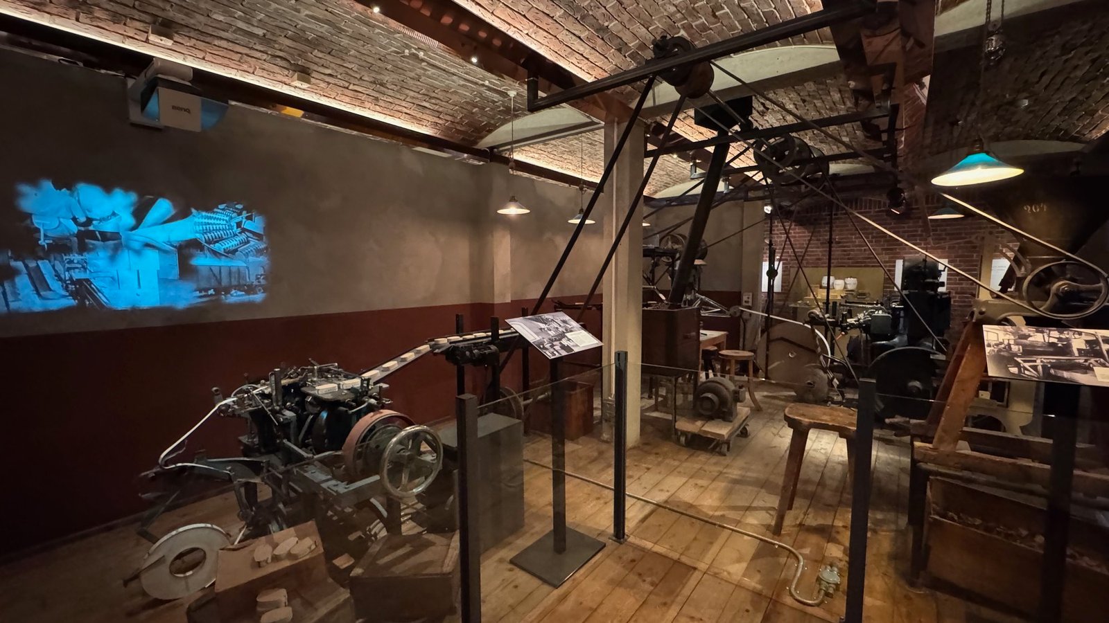 Old factory machines with belts and wheels on a wooden floor in a hall with a brick vault ceiling, and a film projected in blue light on the wall.
