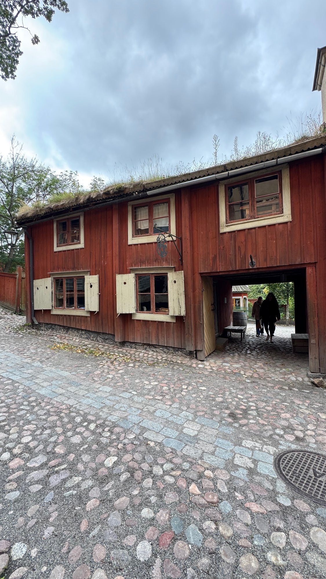 Red wooden house with cream window frames, open cream shutters and a grass roof beside a cobbled lane, with a low gateway where two visitors walk through.