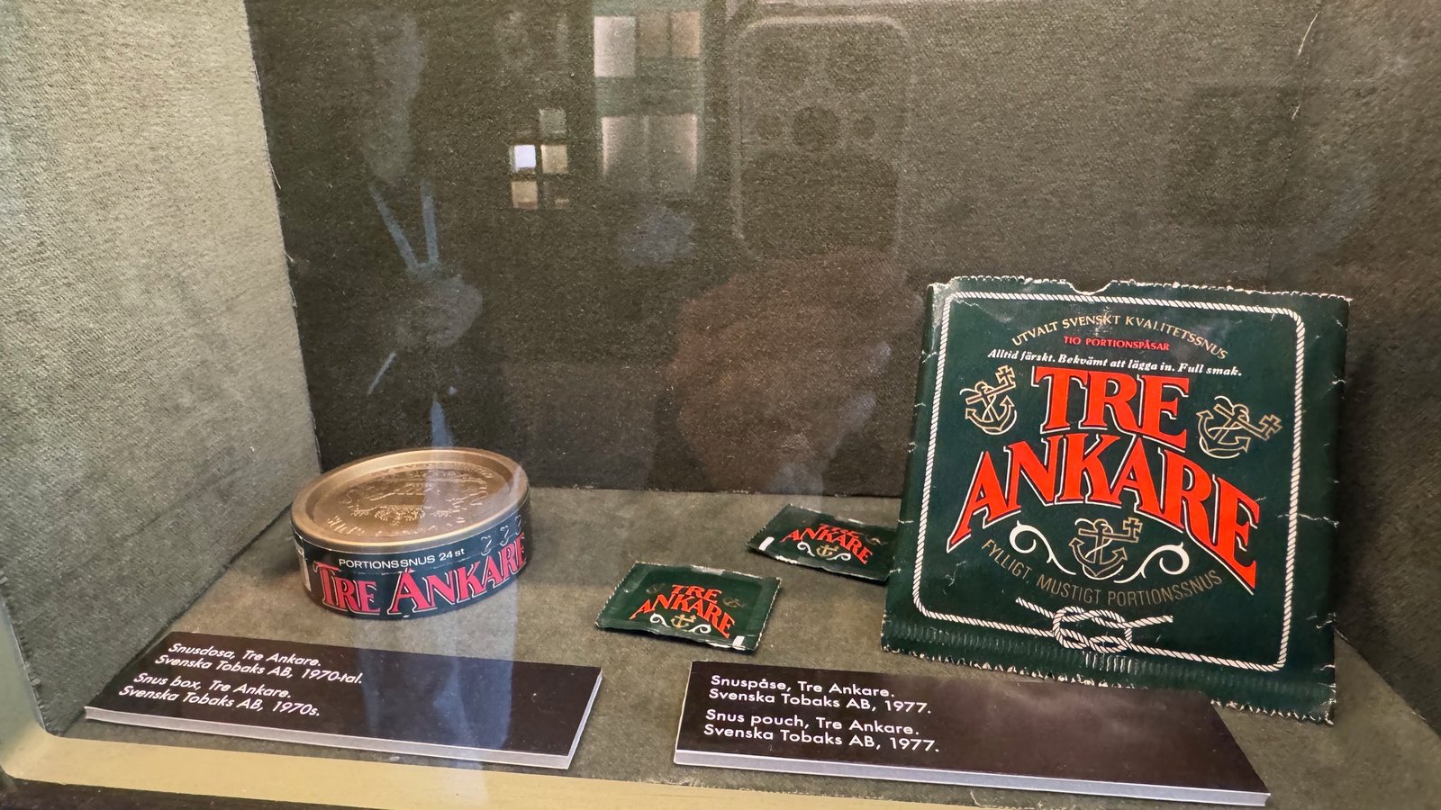Display case with a round Tre Ankare snus tin and green snus pouches with red lettering and anchors, with small labels below.
