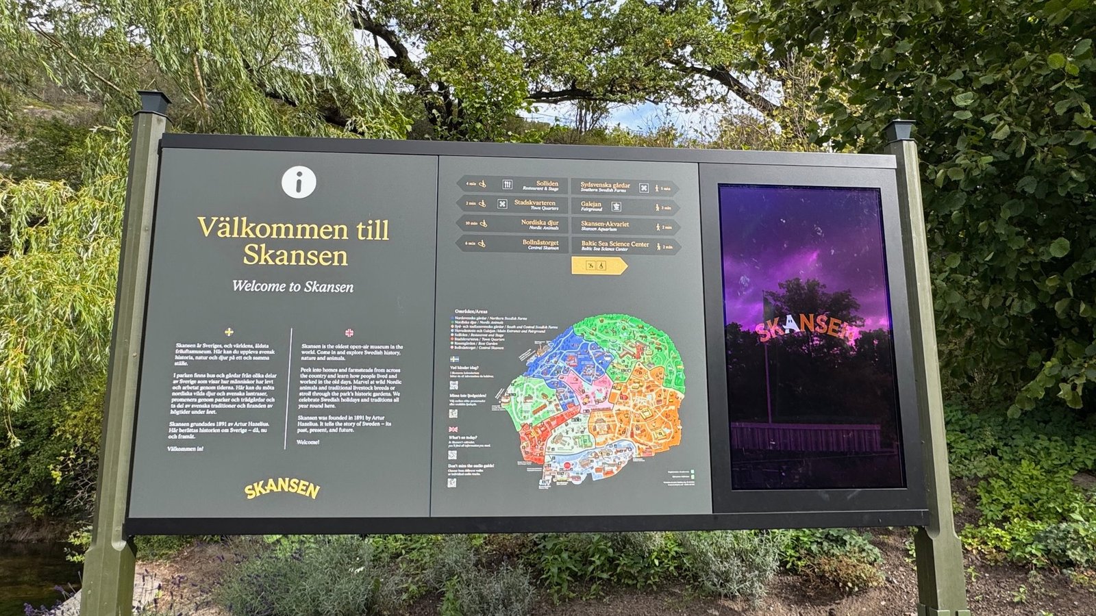Dark welcome sign in front of trees with the text Välkommen till Skansen, a colorful map of the park and a screen showing the word Skansen in orange letters.