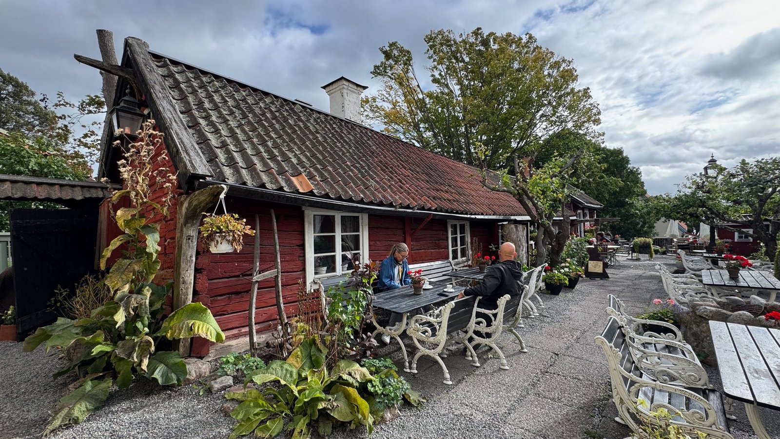 Garden café with tables on a lawn in front of a red log cottage with white window frames, under trees.