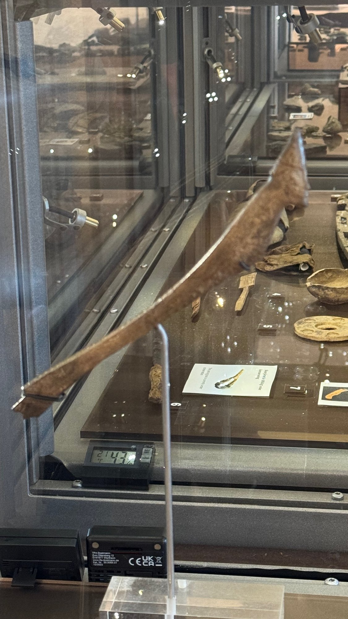 Long narrow curved antler mount in a glass display case. The pointed upper end carries a small carved head of a man in a pointed helmet. Other small finds lie beside it.