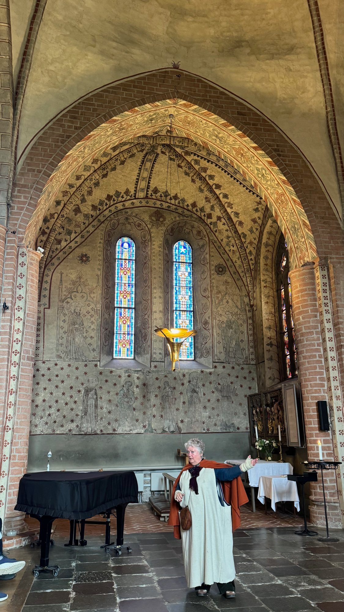 Church choir with painted murals, stained glass windows, a piano and a guide in costume standing at the front.