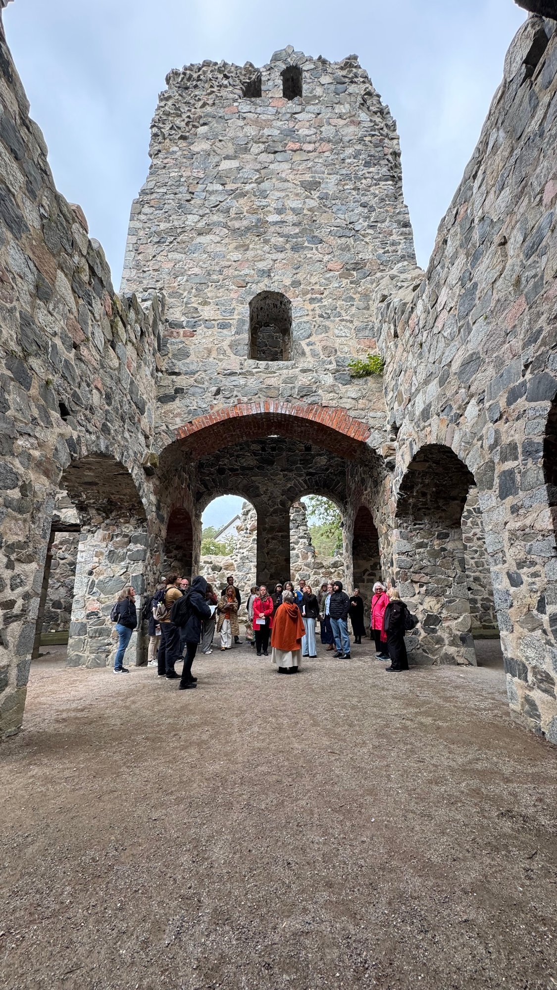 Inside a gray stone church ruin with a tall tower wall and arches open to the sky, a group of visitors and a guide in costume standing on the grass.
