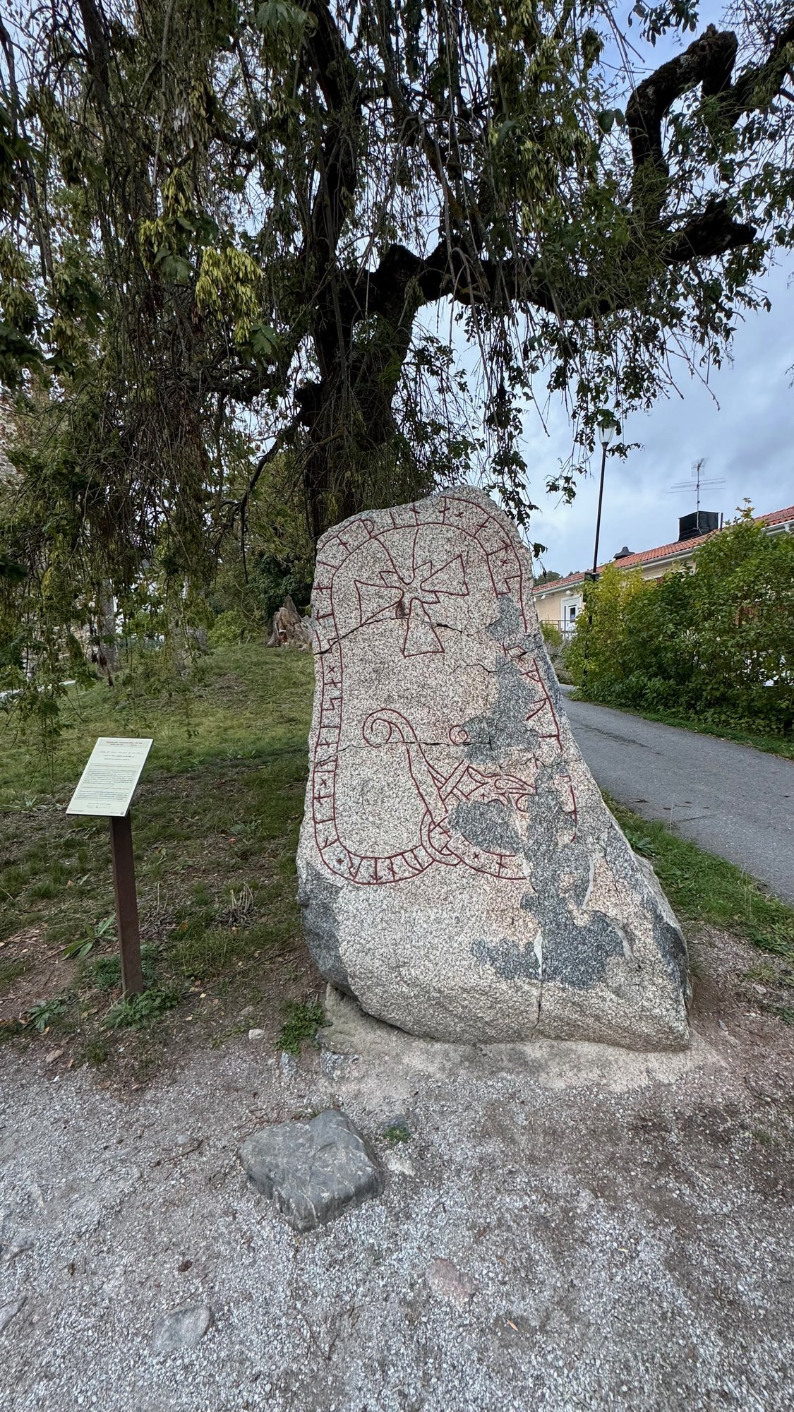 Tall pink granite rune stone with red painted runes, a cross and a serpent band, with an info sign beside it.