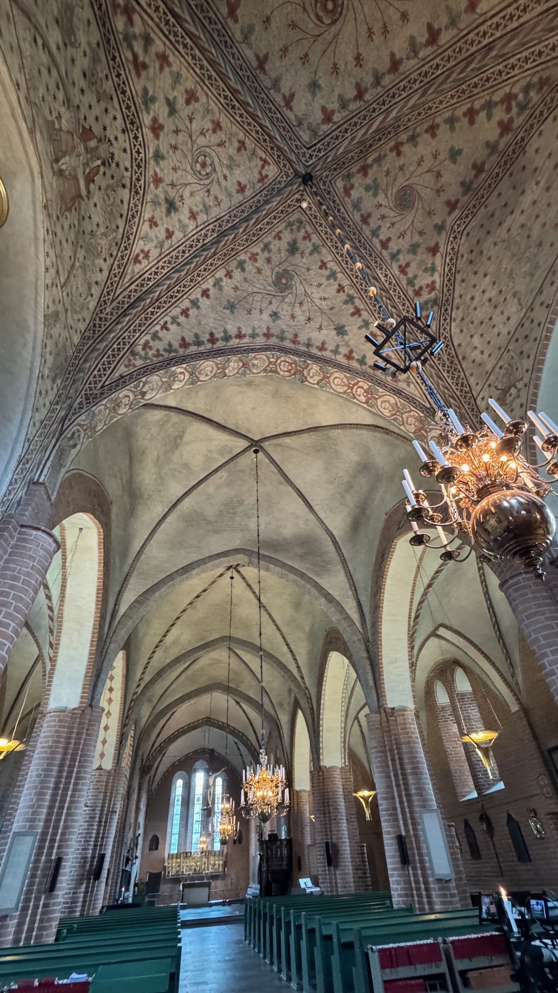 Painted vaulted church ceiling with hanging chandeliers above rows of green pews.
