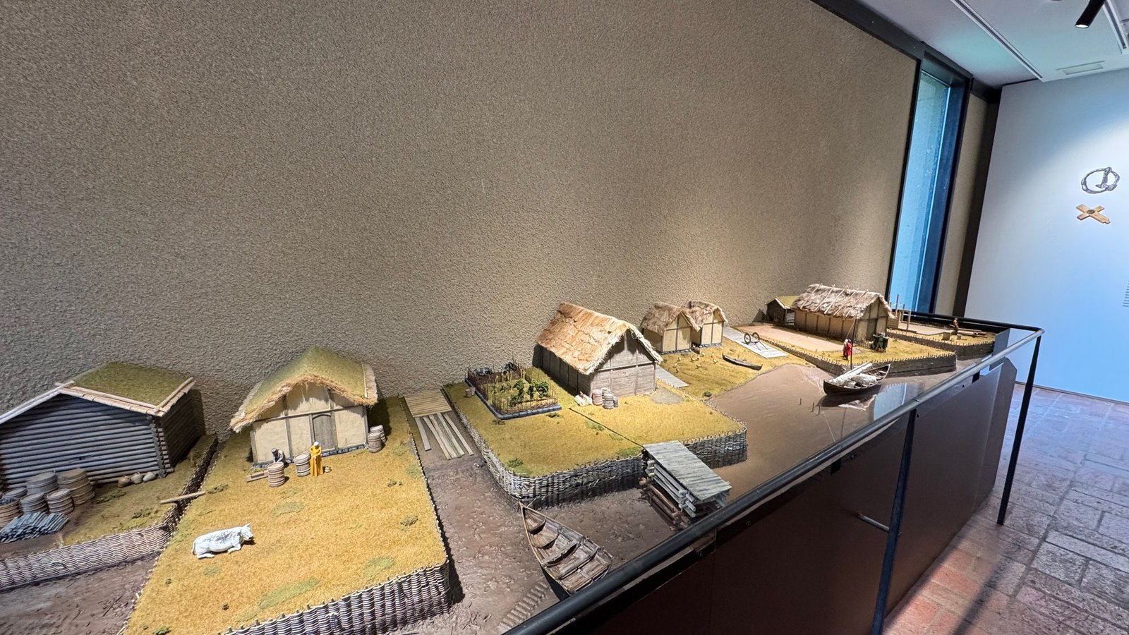 Museum model of Viking Age buildings with small wooden houses, steep roofs and fences on a landscape base, lit in a dark room.