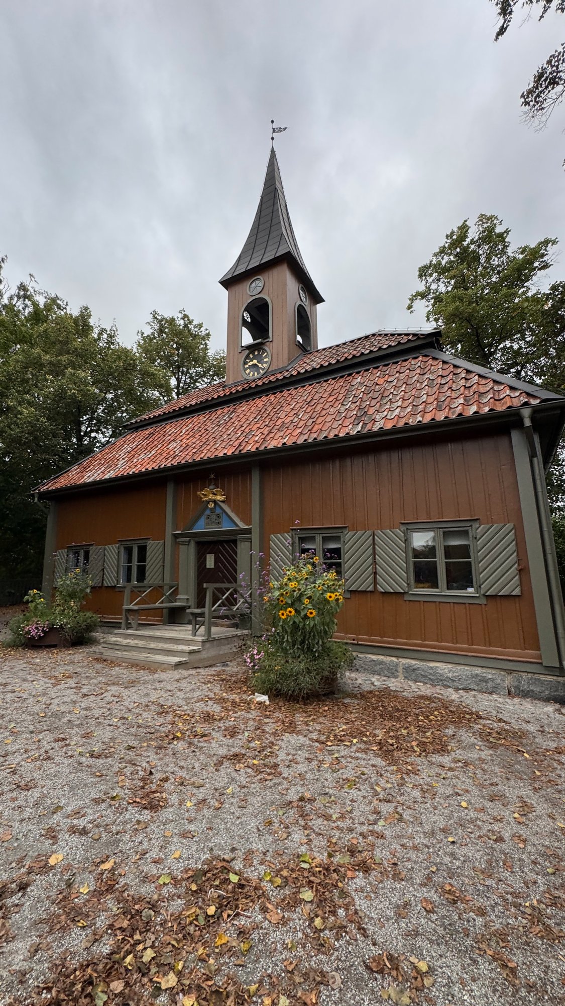 The town hall in Sigtuna, a brown wooden building with a tiled roof, a small clock tower and a crest above the door, beside a cobbled square with a few people.