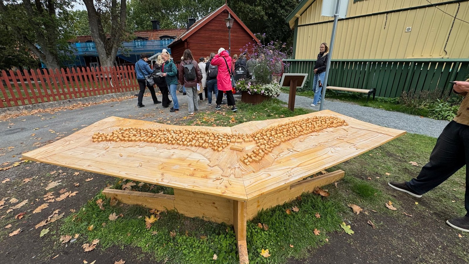 Large wooden model of the town on a stand outdoors, with hundreds of small wooden blocks for houses along two streets, a guided group of visitors behind it and red and yellow wooden houses beyond.