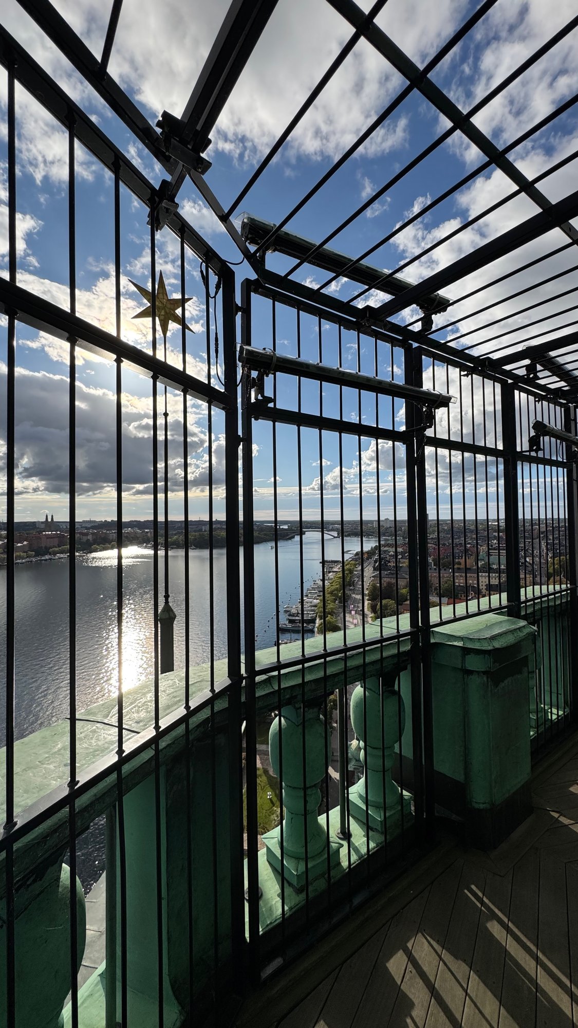 The balcony at the top of the tower with black iron bars, a green copper railing and a small gold star in the corner, with the water and the city behind.