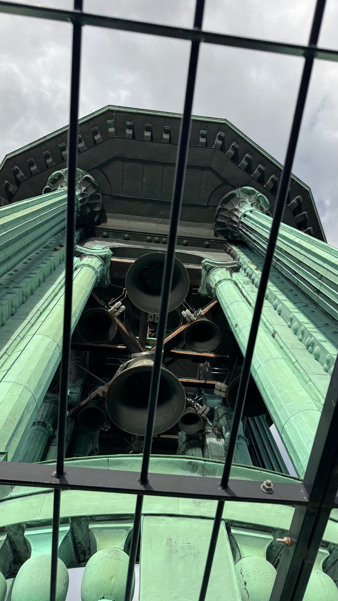 View up through black iron bars at green copper columns and several dark bells in the top of the tower, with grey clouds behind.