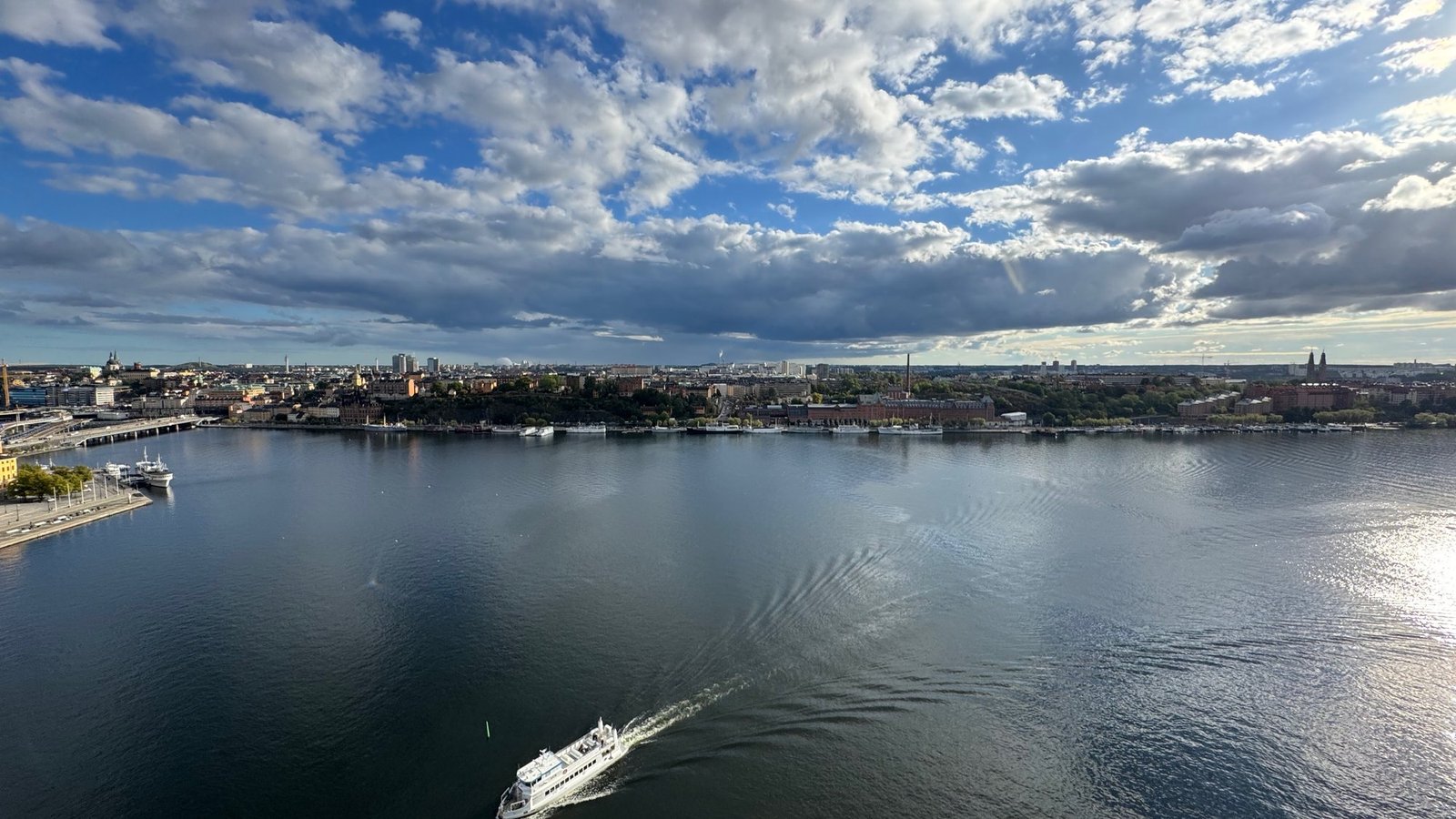 Wide view from the tower over dark water with a white boat and its wake at the bottom, the far shore on the horizon and a sky full of clouds.