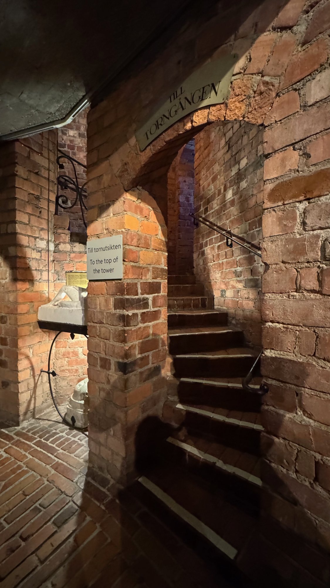 Narrow spiral stair with worn stone steps inside a red brick passage, with a handrail and a small sign that points to the top of the tower.