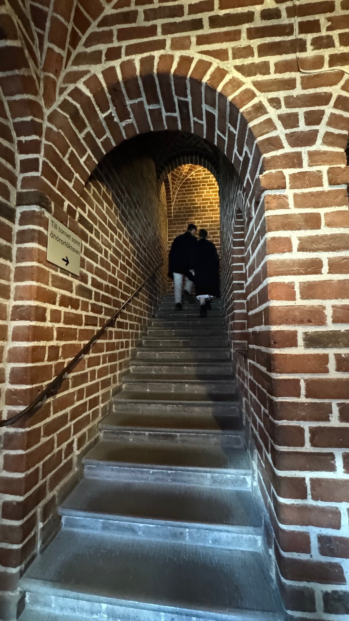 Narrow stairway with stone steps and a handrail between red brick walls and an arched brick vault, with two people at the top.