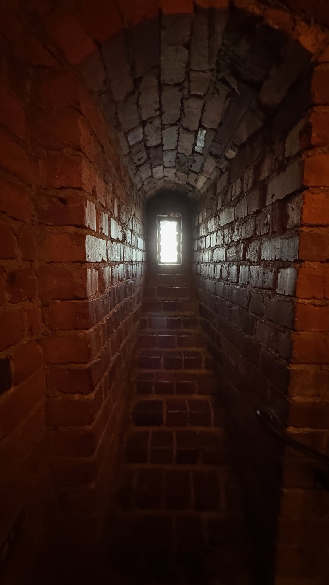 Dark, narrow brick tunnel with a curved ceiling and a steep floor that goes up toward a small bright window at the end.