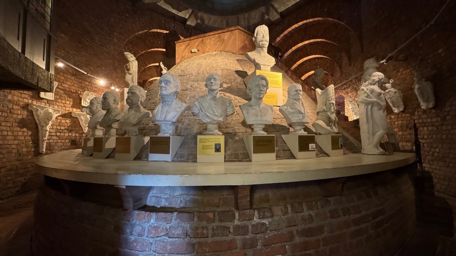 Wide view of a round platform in the tower with a row of white plaster busts with small signs, in front of a brick wall and a vault.