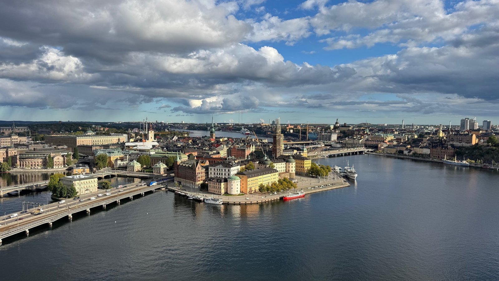 View from high above over dark blue water to the buildings of Gamla stan and Riddarholmen, with a bridge on the left and a big cloudy sky.