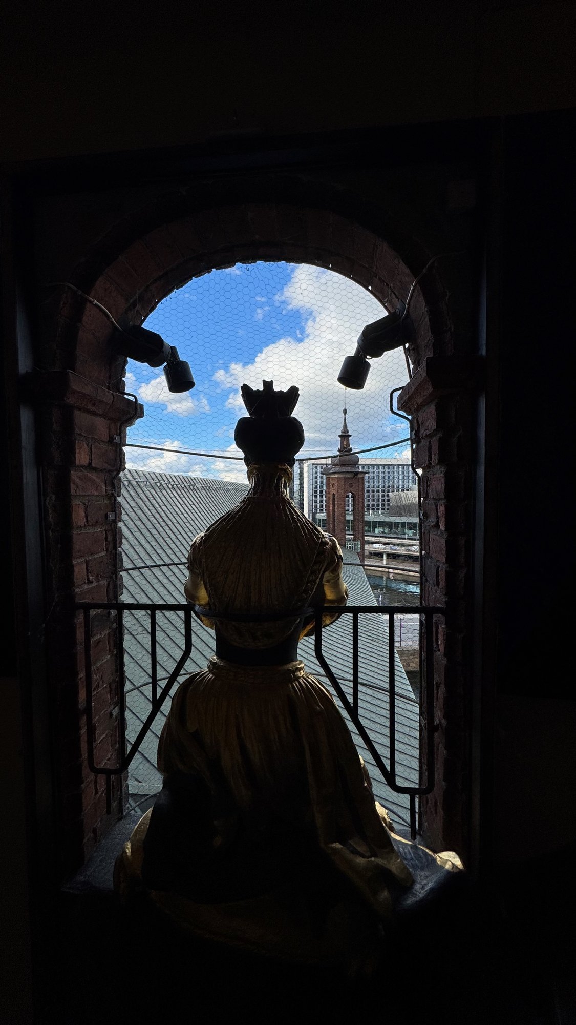A gilded crowned statue seen from behind, in an arched brick window with blue sky and the roofs of the city beyond.