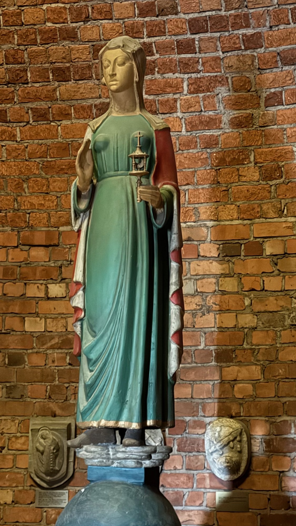 A painted wooden statue of a standing woman in a green dress with a red cloak, on a round base in front of a red brick wall.