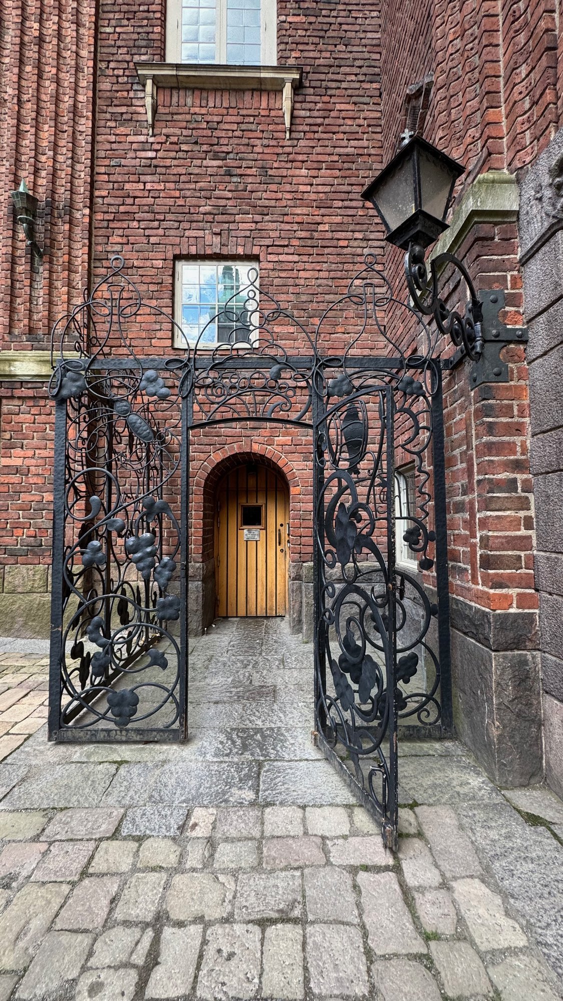 Black wrought iron gate with a lantern in front of a small arched wooden door in a red brick wall, on a cobblestone path.