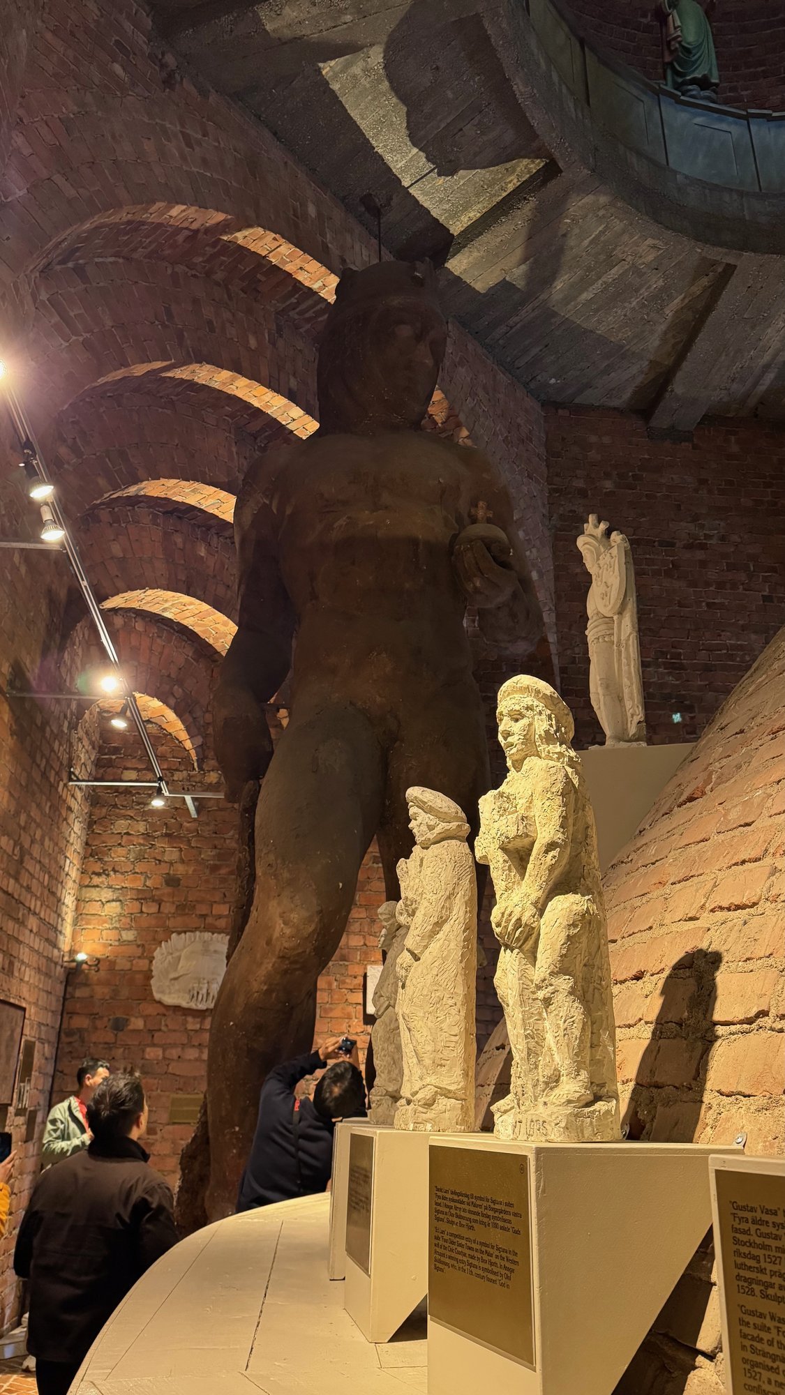 A large dark standing figure against a brick wall with white statues of two figures in front, and curved brick arches in the roof. Some visitors stand at the bottom left.