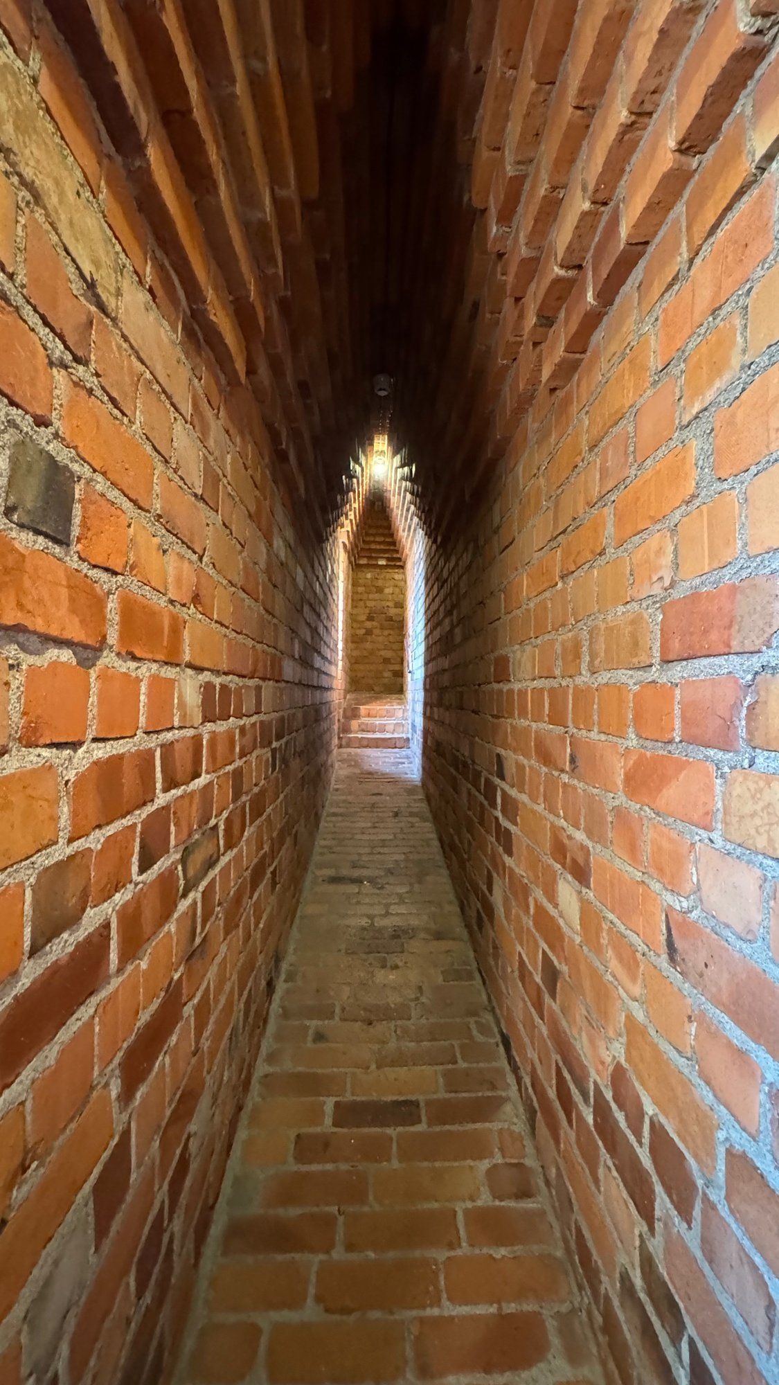 Very narrow passage with orange brick walls and a pointed brick vault, and a pointed opening of light at the far end.