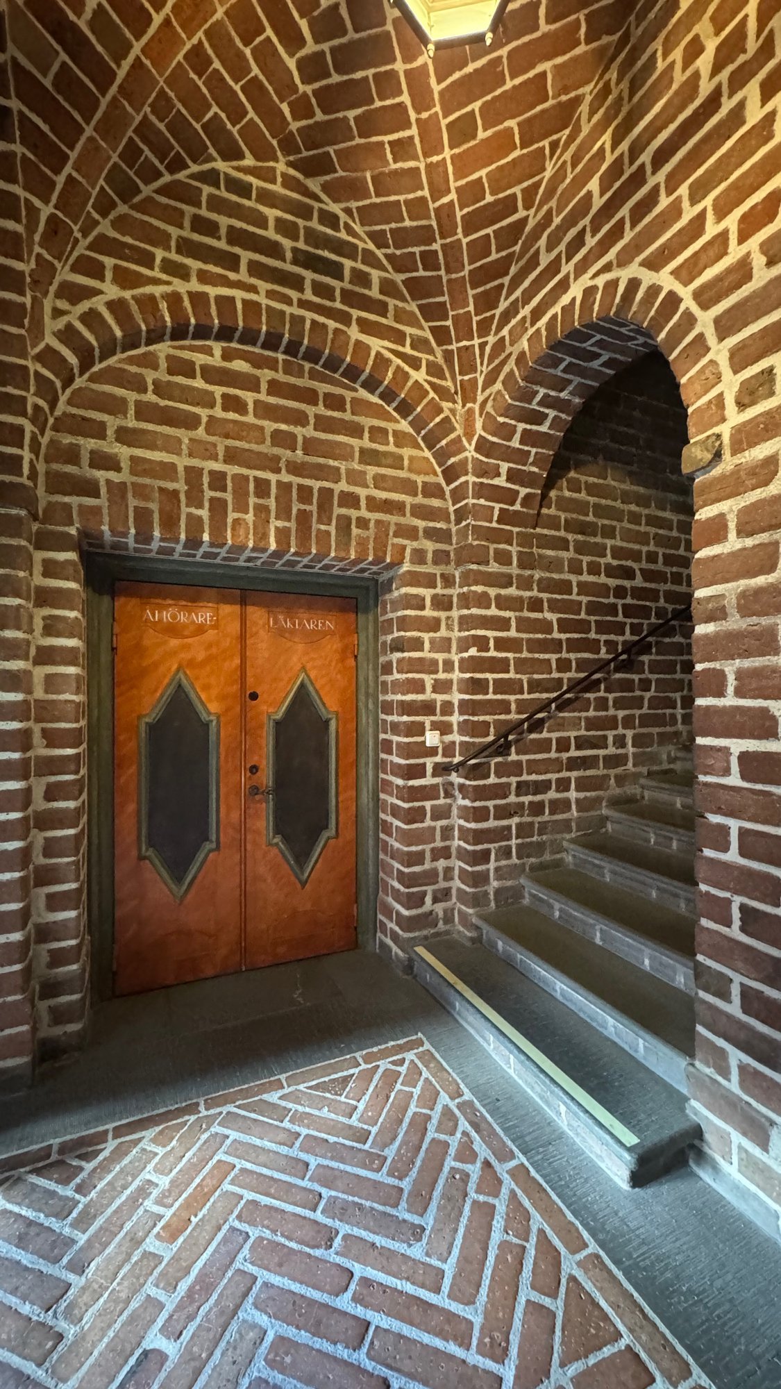 Pair of orange wooden doors with diamond shaped panels under a red brick vault, with a herringbone brick floor and stairs on the right.