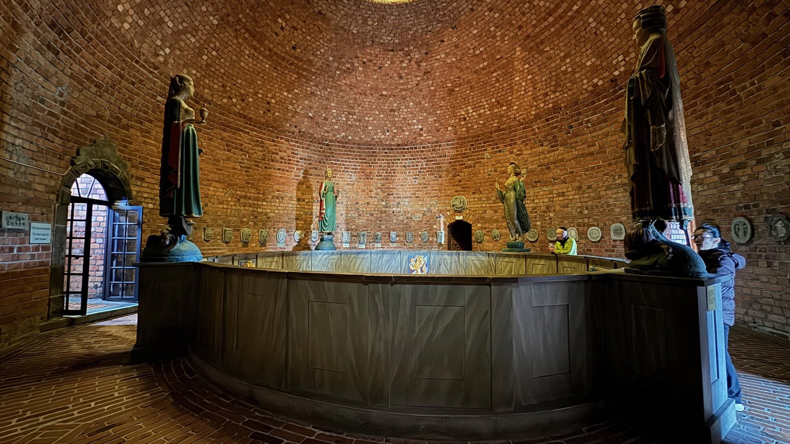 Round platform with a low wooden wall and tall painted statues on it, under a domed brick roof with a wide window on the left.