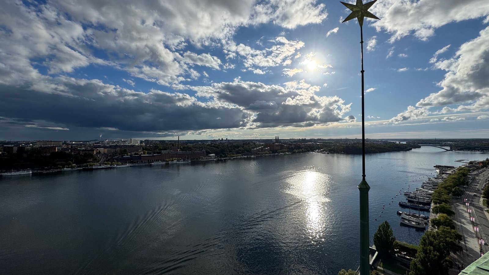 Wide view over dark water with the sun low behind clouds and a bright reflection, the gold star at top right and a quay with boats on the right.