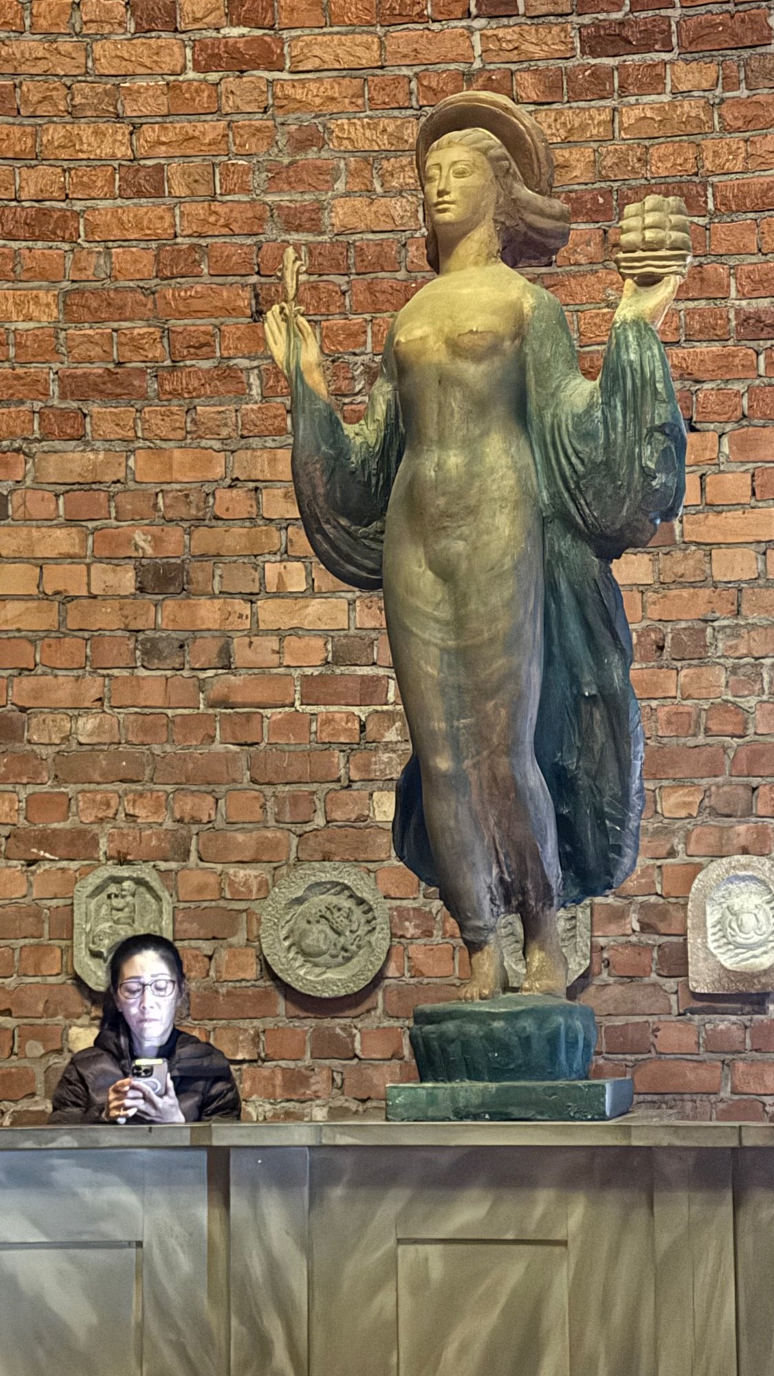 A tall statue of a woman with a raised hand and a green cloak, with round reliefs on both sides, in front of a brick wall. A visitor looks at a phone at the base.