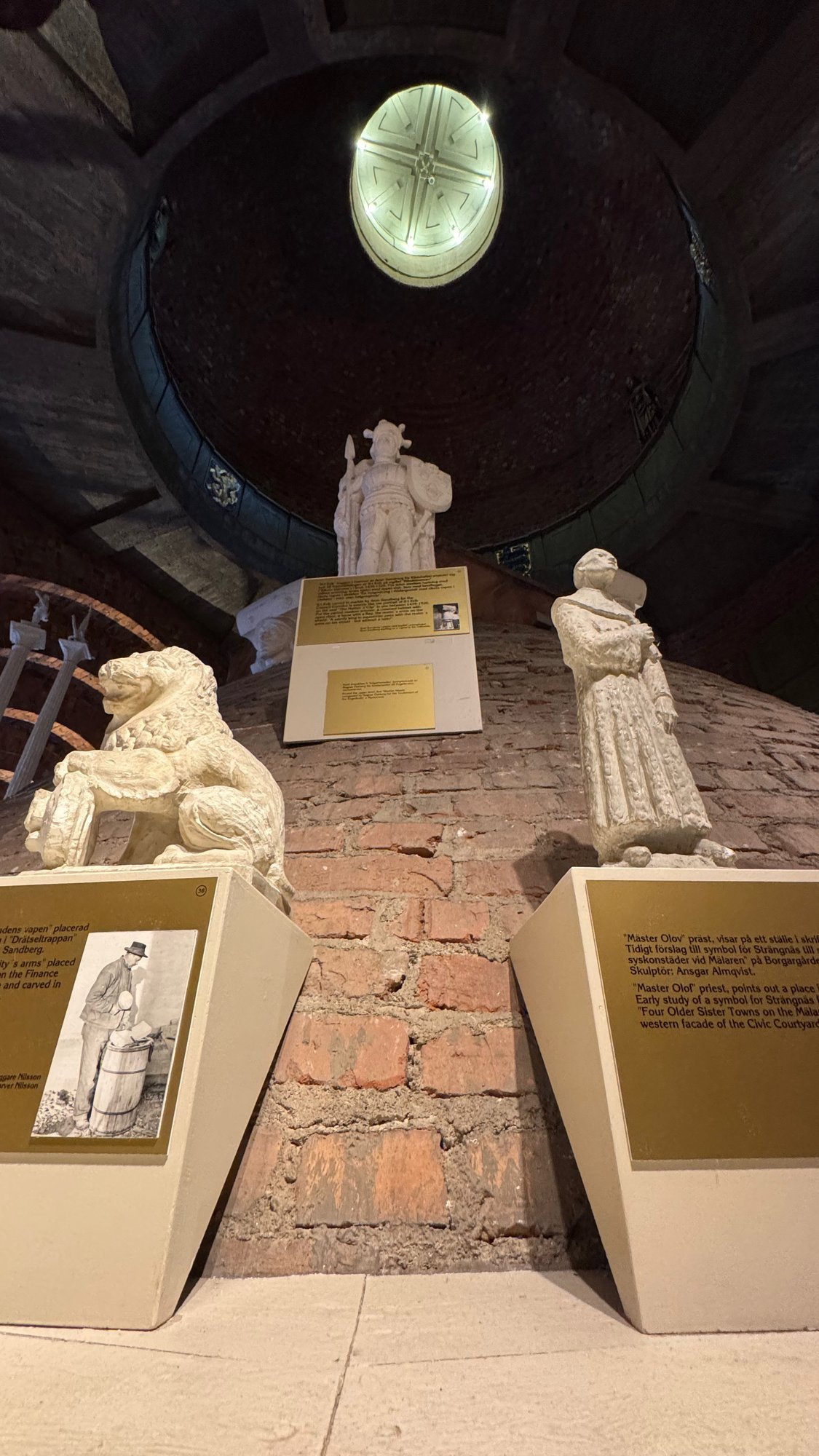 White statues of a lion and two standing figures on a brick slope, with a round lit opening in the dark dome above and brass signs in front.