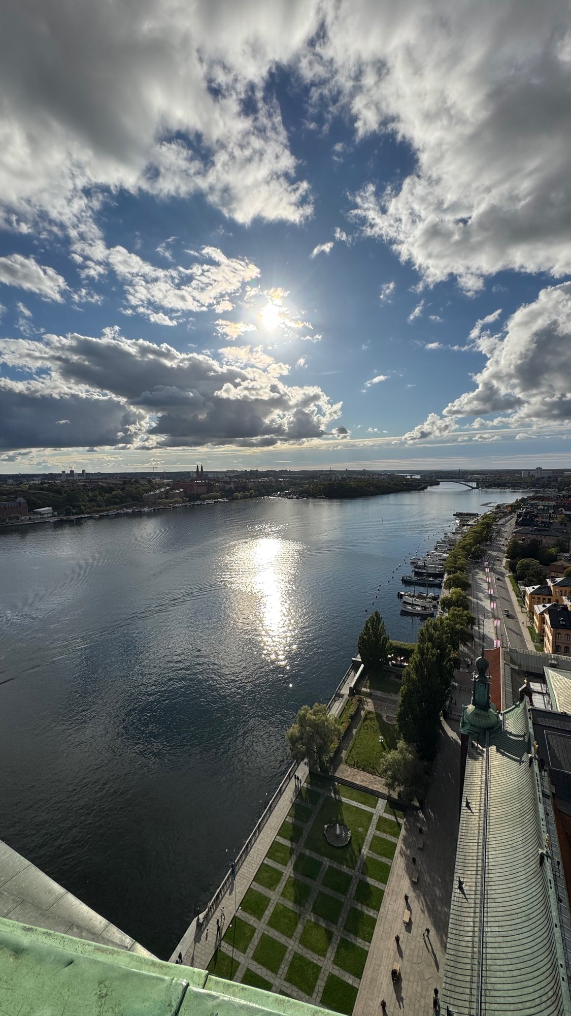View from the tower over the water with a bright sun reflection, a green lawn and a green copper roof at the bottom right, and large clouds above.