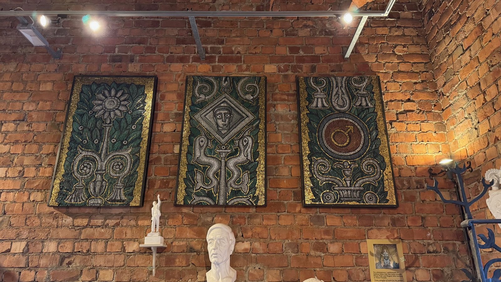 Three tall panels with green and gold patterns and faces, hung on a red brick wall, with a small white statue and a bust below.