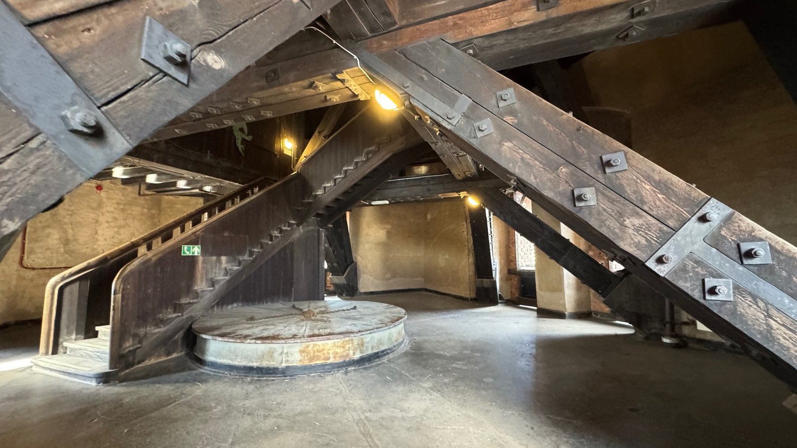 Heavy dark timber beams with iron bolts in a cross over a round metal platform and a wooden stair, in a large room with a concrete floor.