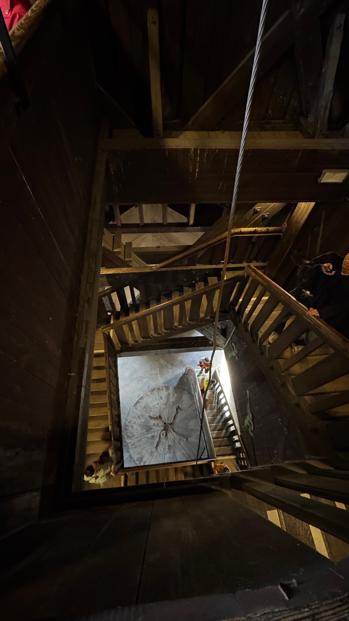 View down a square shaft of wooden stairs with a pale round plate at the bottom, between dark timber beams.