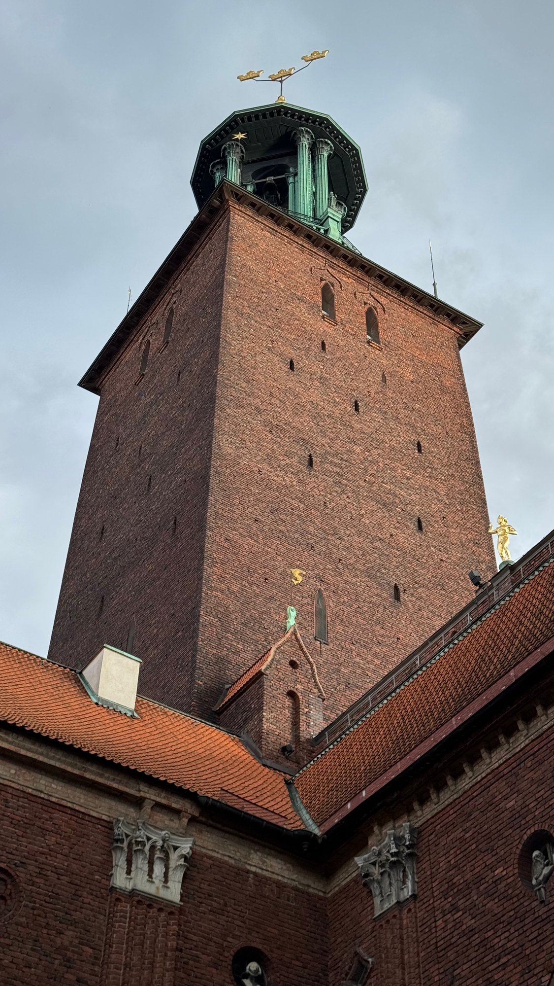 Tall red brick tower with a green copper top and gold crowns, seen from below between brick walls and red tile roofs in the courtyard.