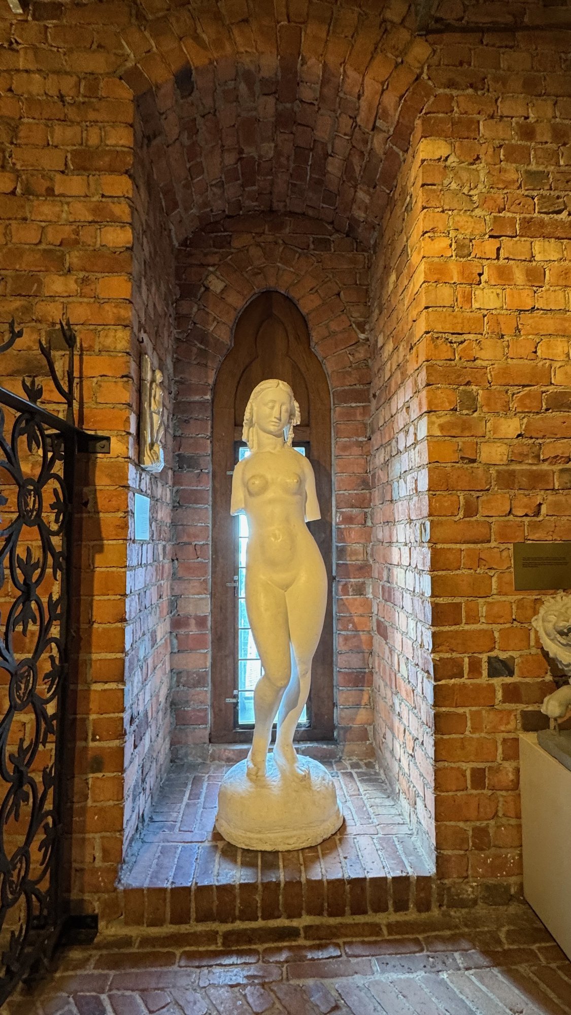A white statue of a standing woman in a narrow brick window niche with an arched top, lit warm from the left.