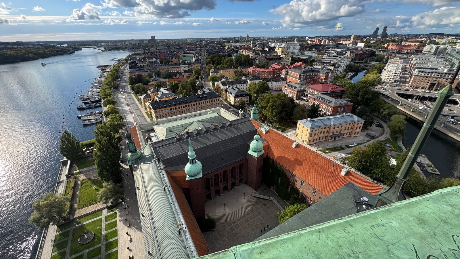 Wide view from the tower over a long brick building with a green roof, the lake and the bright water, with the city and bridges on the right.