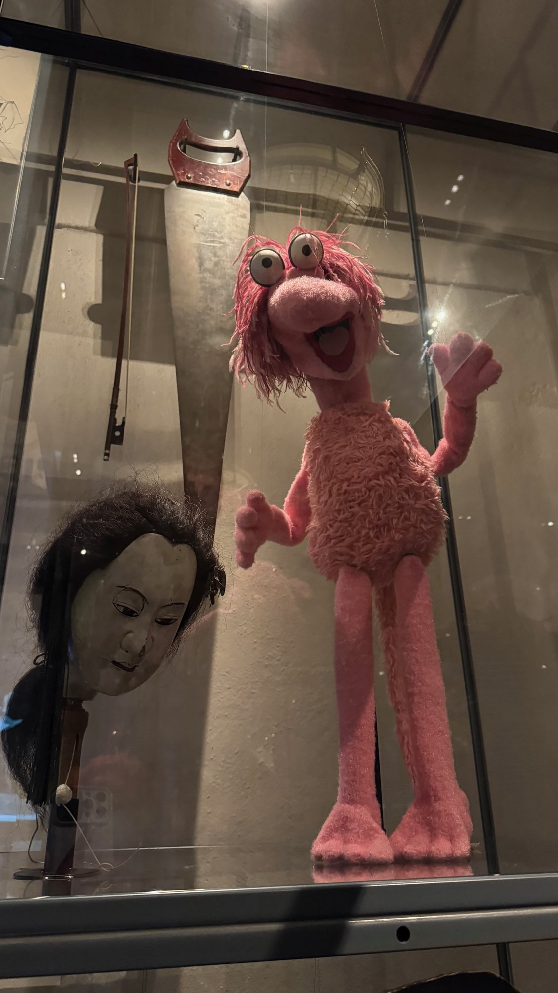 A pink furry puppet and a puppet head in a glass case at Scenkonstmuseet.