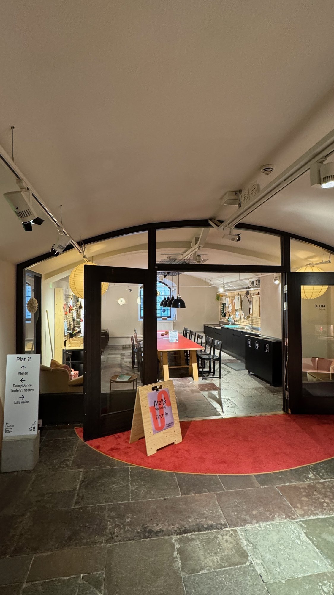 Open glass doors with a red carpet lead into the Studio at Scenkonstmuseet, with a red worktable, chairs and a sign that says Ateljé, drop in.